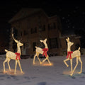 Three gold lighted 2D deer with red bows standing on a snowy lawn at night