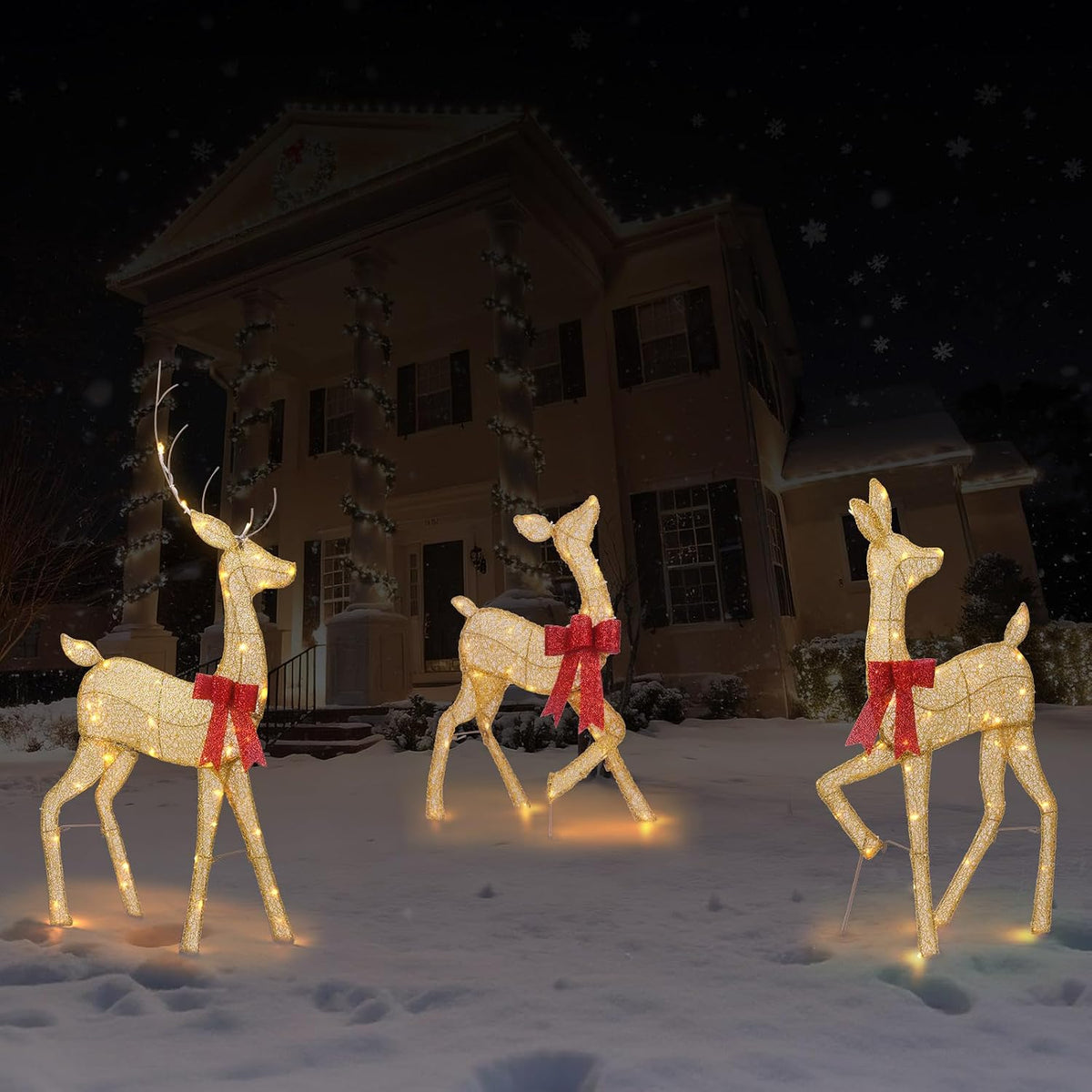 Three gold lighted 2D deer with red bows standing on a snowy lawn at night