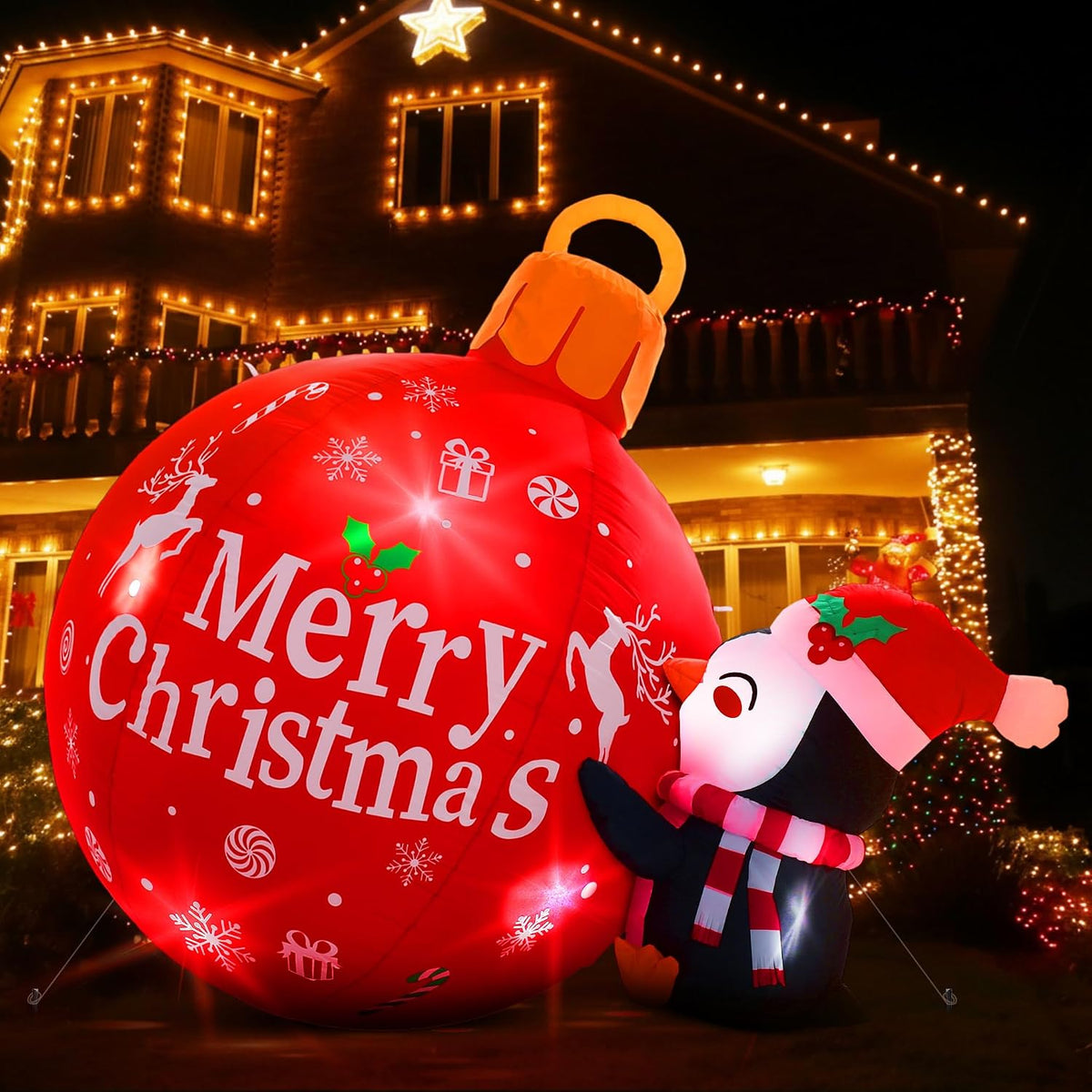 Penguin in a Santa hat beside a glowing red Merry Christmas ornament inflatable in front of a lit house